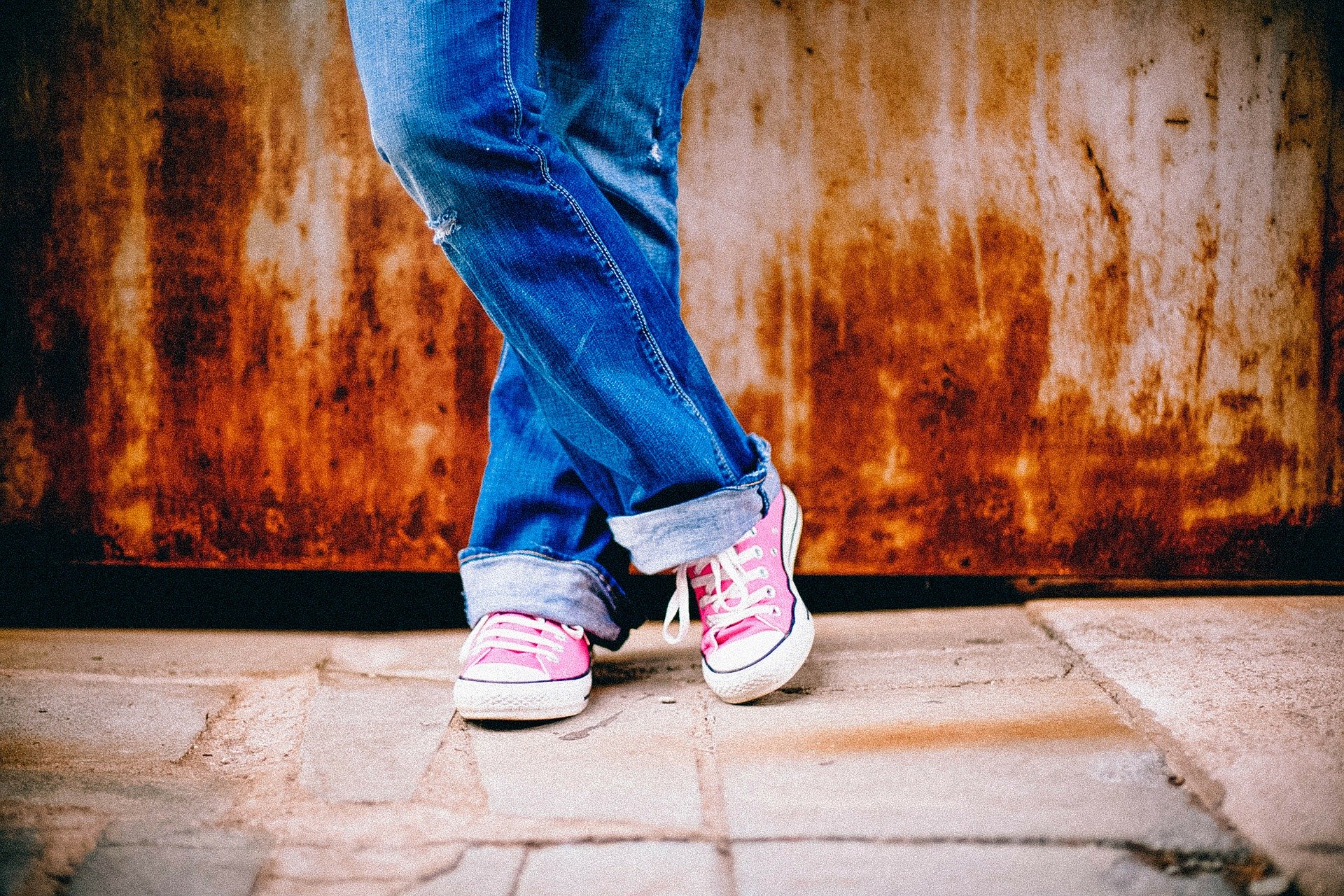 person's legs standing in front of a rusty wall, legs crossed, wearing blue jeans and pink and white sneakers